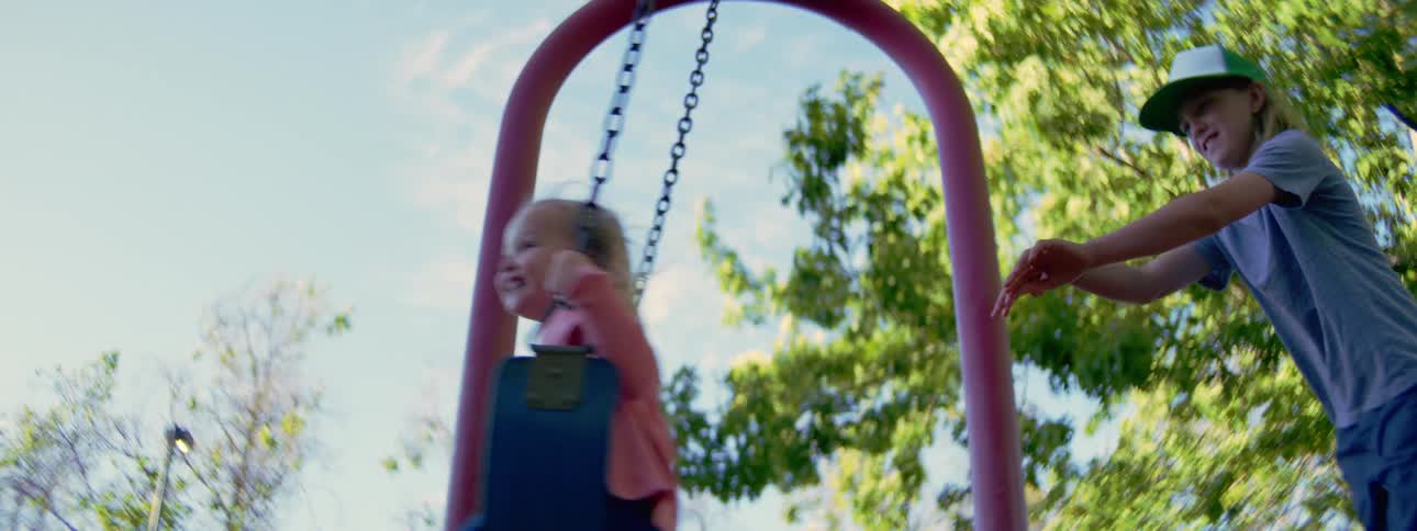 In slow motion, a big brother playfully pushes his laughing little sister on a swing set at the park
