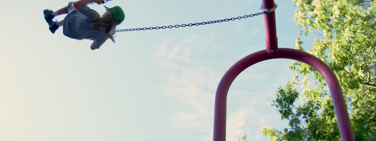 From a low angle perspective, a child in a green hat swings back and forth in slow motion on a sunny day