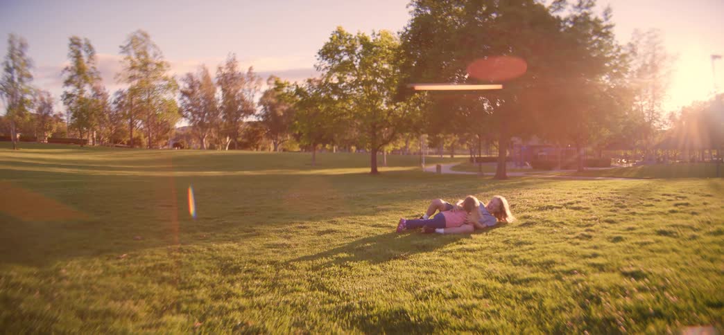 A young girl runs across a sunlit park to her older brother, and they playfully fall into the grass together in a hug, filmed in slow motion