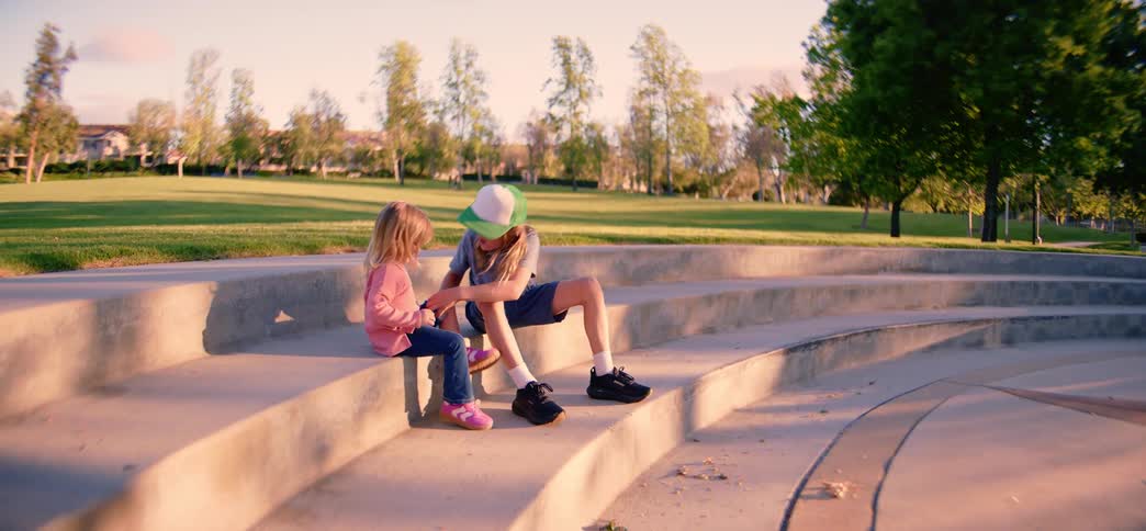 An older brother helps his little sister with her shoe as they sit together on concrete amphitheater steps in a park at golden hour