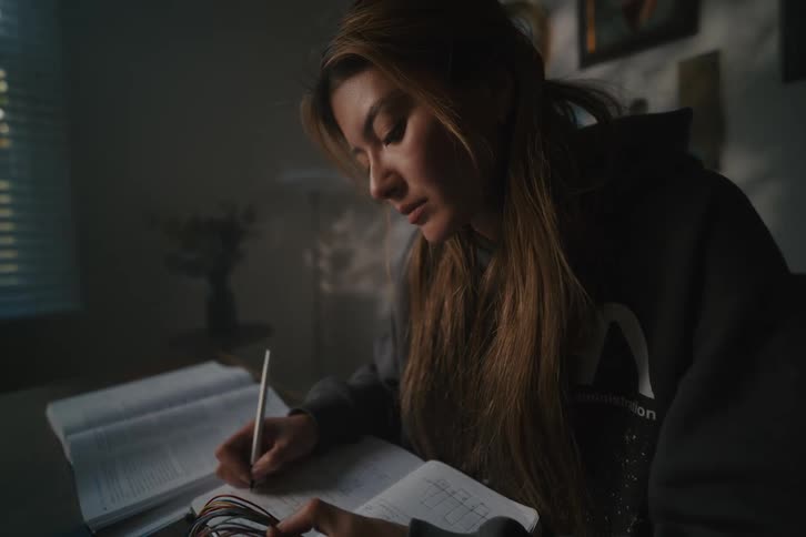 A focused young woman studies in a dimly lit room, diligently writing notes from her textbook late at night