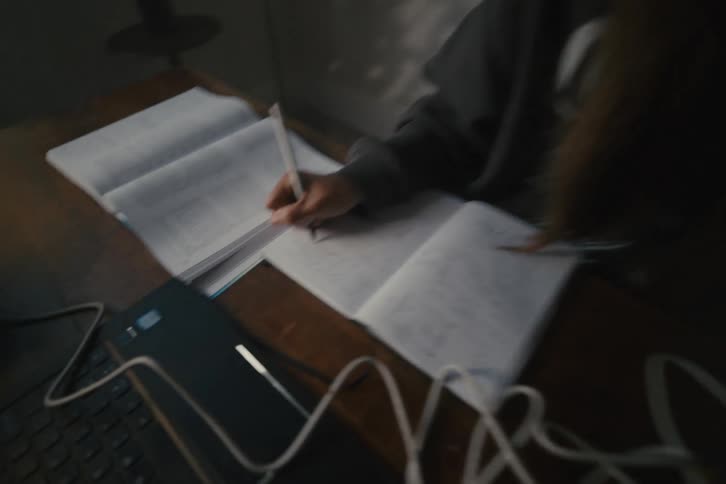 A focused female student diligently writes in her notebook while studying from a textbook and laptop in a dimly lit room