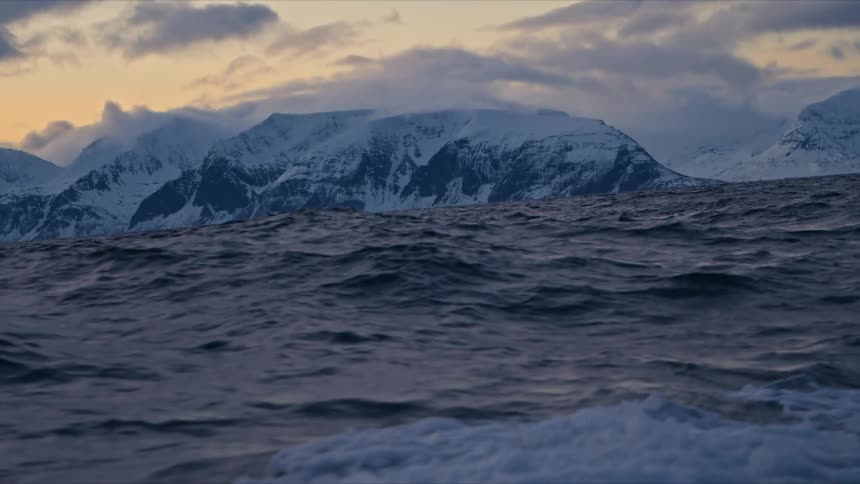 Choppy dark blue ocean waves move past the camera with majestic snow-covered mountains in the distance under a warm, cloudy sky