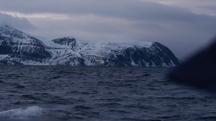 Slow-motion shot from a boat on a choppy sea, showing a massive snow-covered mountain under a moody, overcast sky