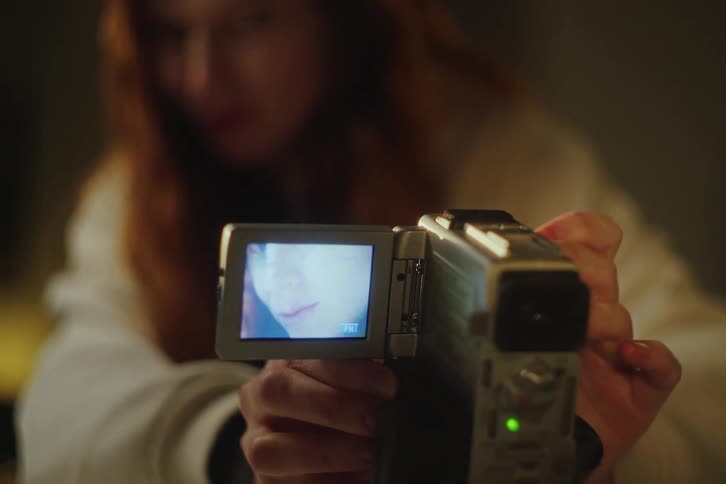 A woman holds a vintage digital camcorder, filming a self-portrait that's visible on the flip-out screen