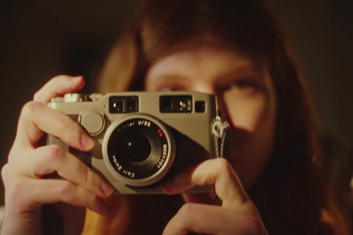 A close-up of a young woman with red hair taking a picture with a vintage film camera in a dimly lit room