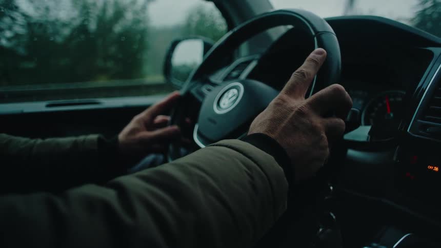 A moody close-up shot shows a man's hands firmly on the steering wheel while driving on a rainy day