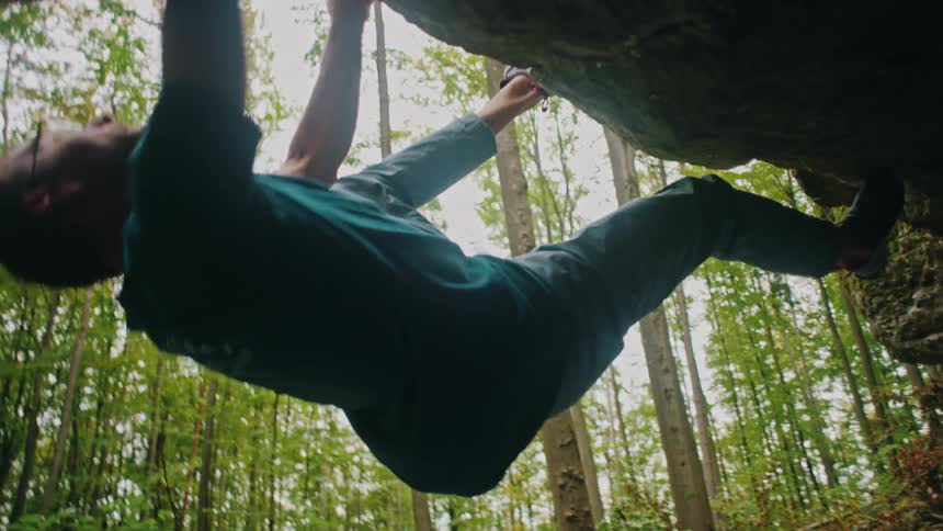 From a low angle, a strong rock climber traverses an overhang in a green forest before losing his grip and falling