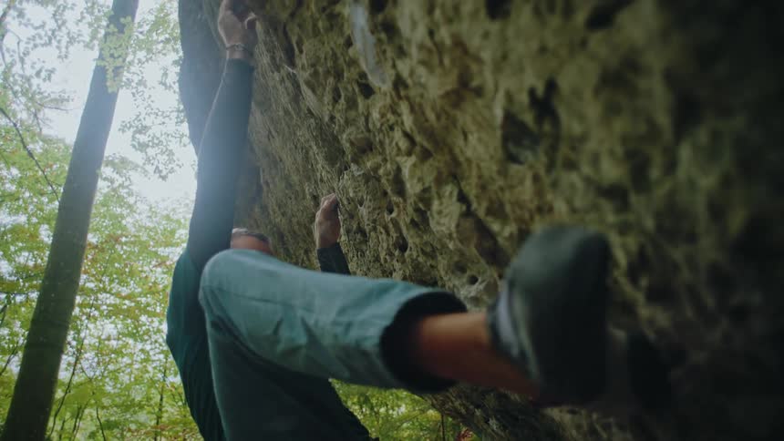 A focused climber in slow motion skillfully navigates a difficult bouldering route on an overhanging rock in a dense forest