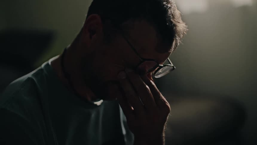 A close up profile shot of a distraught man with glasses rubbing his tired eyes in a dimly lit room