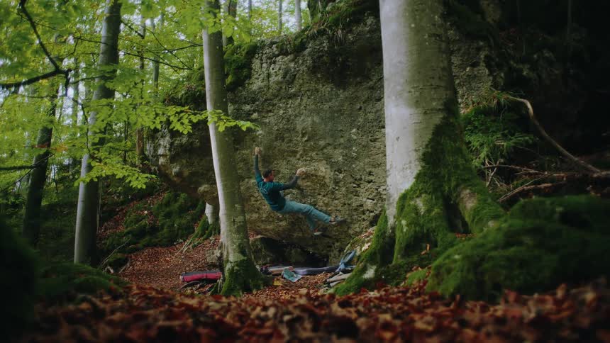 A climber in slow motion meticulously scales a large boulder in a serene, moss-covered autumn forest