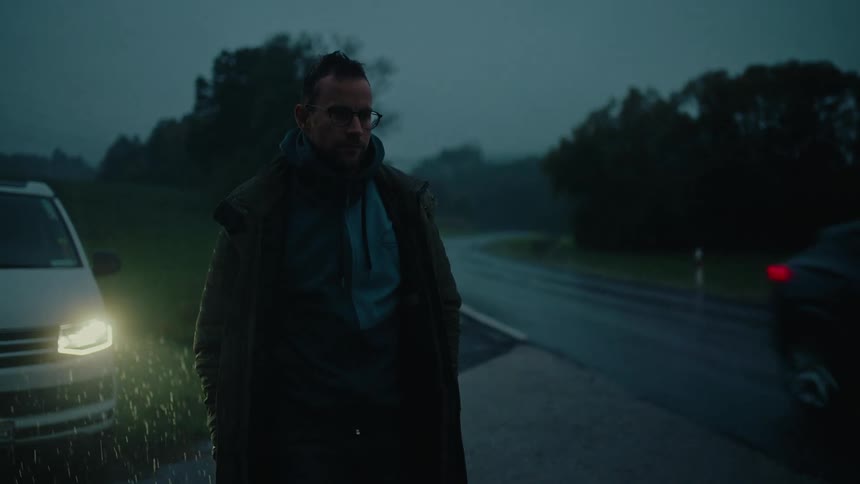 A lone man waits in the pouring rain on the side of a dark, rural road at night while a car passes by