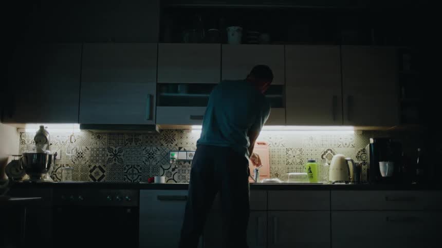 A man from behind washes dishes in a modern, dimly lit kitchen at night