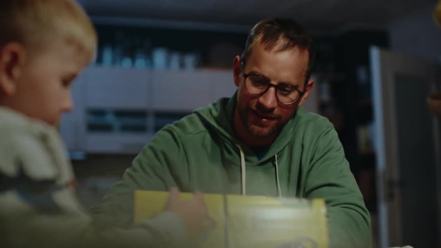 Caring father and his young son bond while looking through an educational book together at the table in the evening