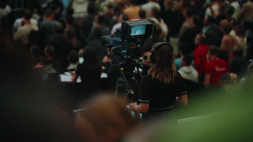 A female camera operator, seen from behind, films a large crowd at an indoor conference or event
