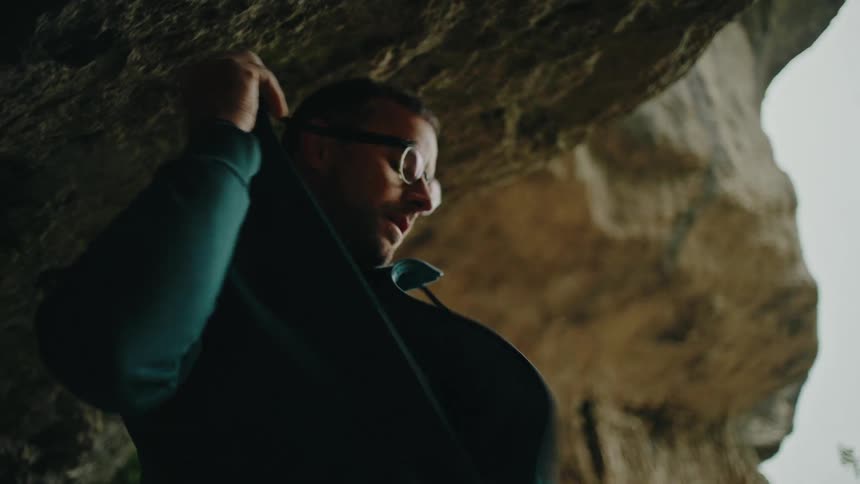 Slow motion shot of a man with glasses taking off his green hoodie inside a dark, rocky cave