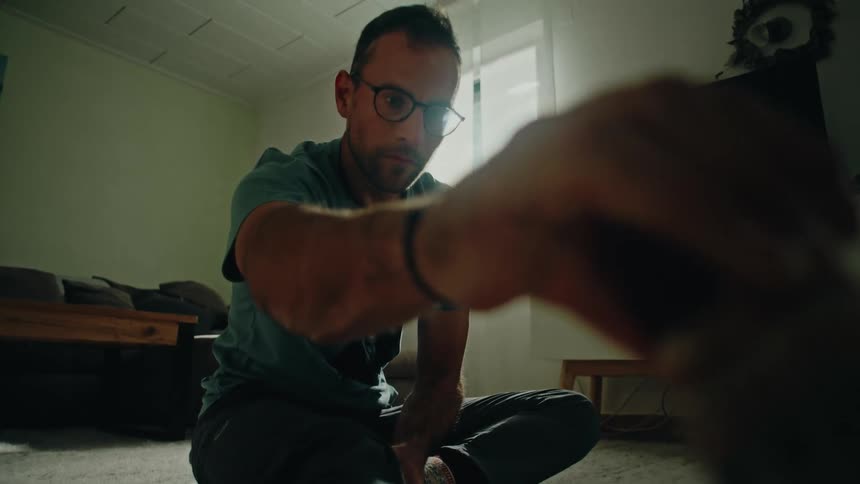 A man in glasses stretches his leg while sitting on the floor of his living room, shot from a low angle