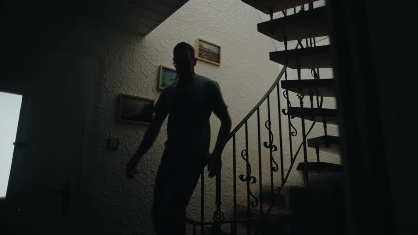 A silhouetted man in glasses slowly walks down a winding spiral staircase in a dark, atmospheric home