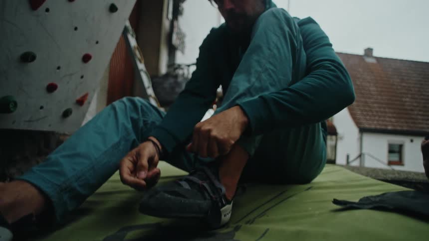 A man in a beanie and glasses sits on a crash pad and puts on his climbing shoes in slow motion next to an outdoor bouldering wall