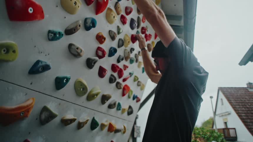 A focused man in a beanie diligently trains on a colorful outdoor bouldering wall in slow motion