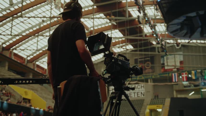 A professional camera operator records a live event from a raised platform inside a crowded international arena