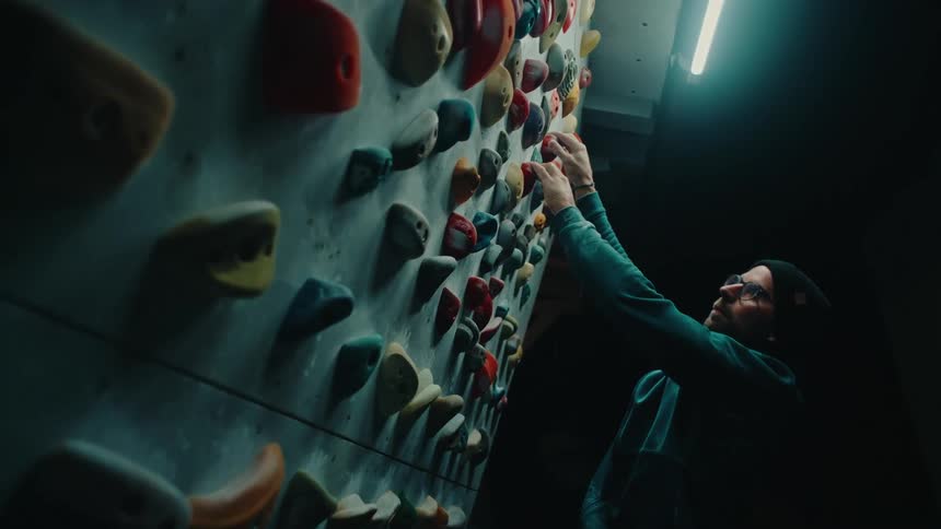 A man in a beanie and glasses carefully ascends an indoor bouldering wall in a dark, atmospheric gym in slow motion