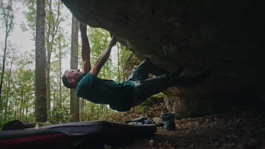A strong male rock climber navigates a challenging overhang boulder problem in a forest, ultimately falling onto the crash pad below in slow motion