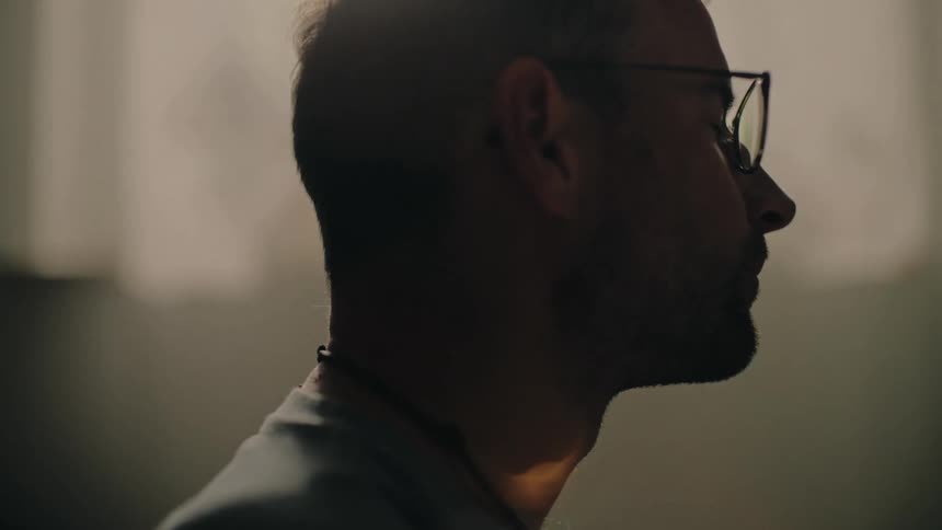A stressed man wearing glasses sighs and rubs his eyes in a moody, backlit room