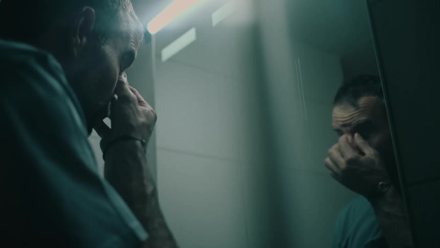 A troubled man confronts his reflection in a dimly lit bathroom, rubbing his tired face in despair