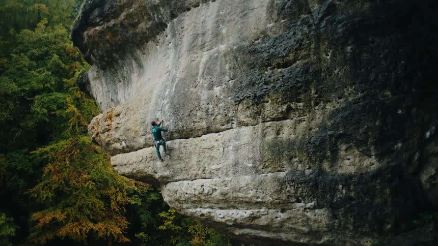 A lone rock climber ascends a challenging cliff face surrounded by a lush forest
