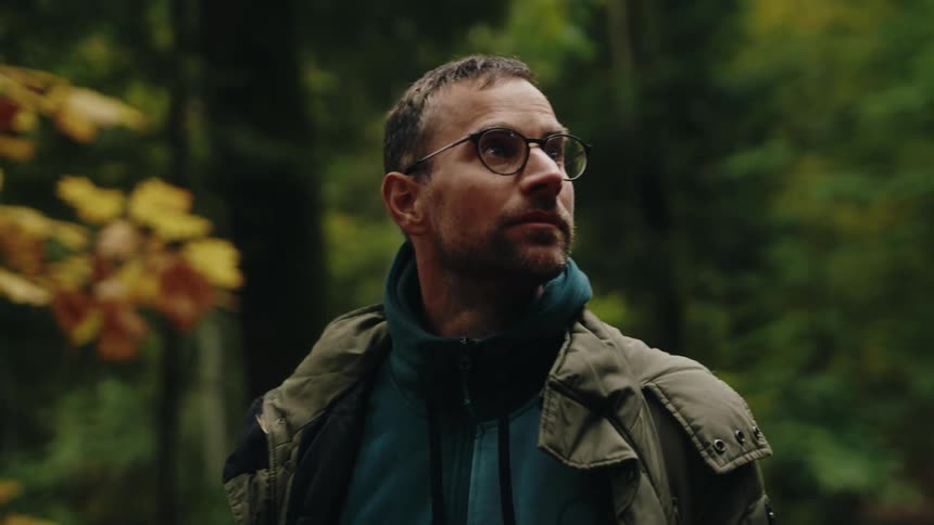 A contemplative man wearing glasses and a jacket pauses to admire the surrounding nature while hiking in a tranquil autumn forest