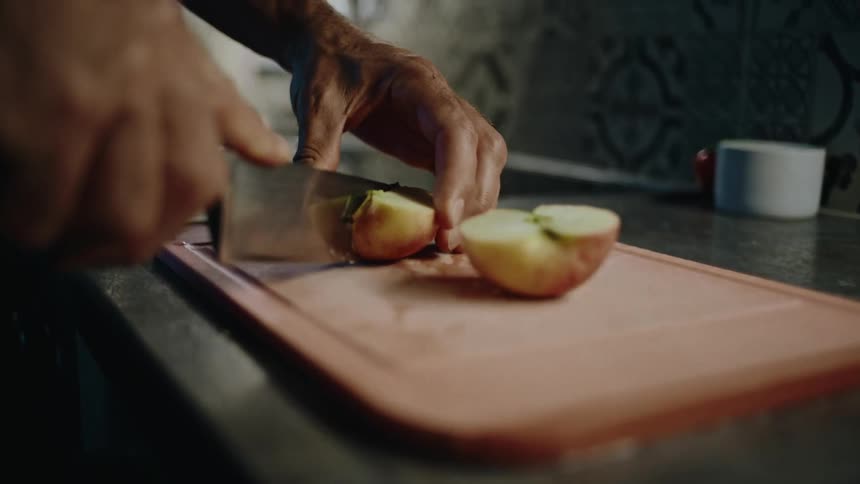 A man's hands slowly slice a fresh apple with a knife on a cutting board in a dimly lit kitchen