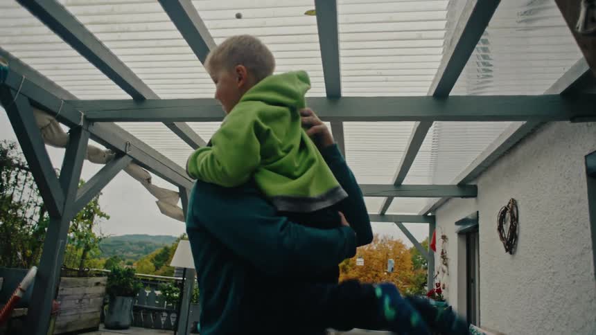 In slow motion, a loving father playfully lifts his young son and holds him in a warm embrace on their home patio