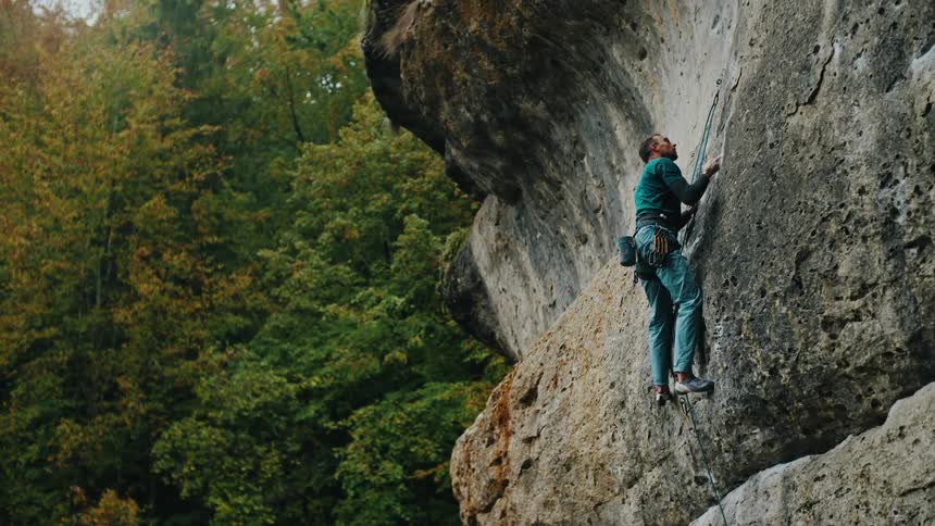 A determined rock climber carefully ascends a steep cliff face in a dense, green forest