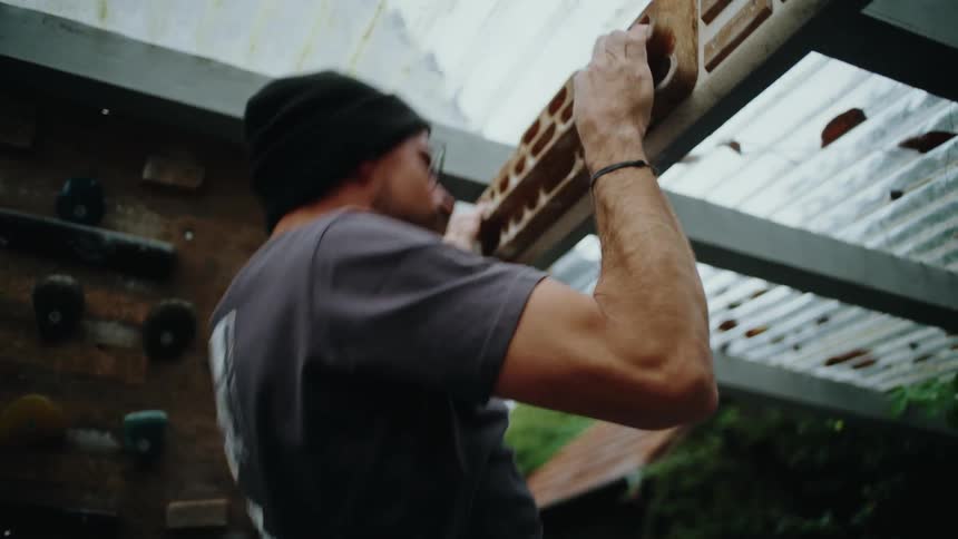 A determined man in a beanie does pull-ups on a wooden hangboard as part of his rock climbing training in a home gym