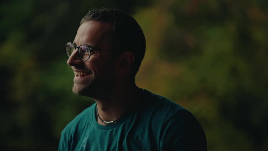 A man wearing glasses and a green shirt smiles and laughs genuinely in a softly lit setting