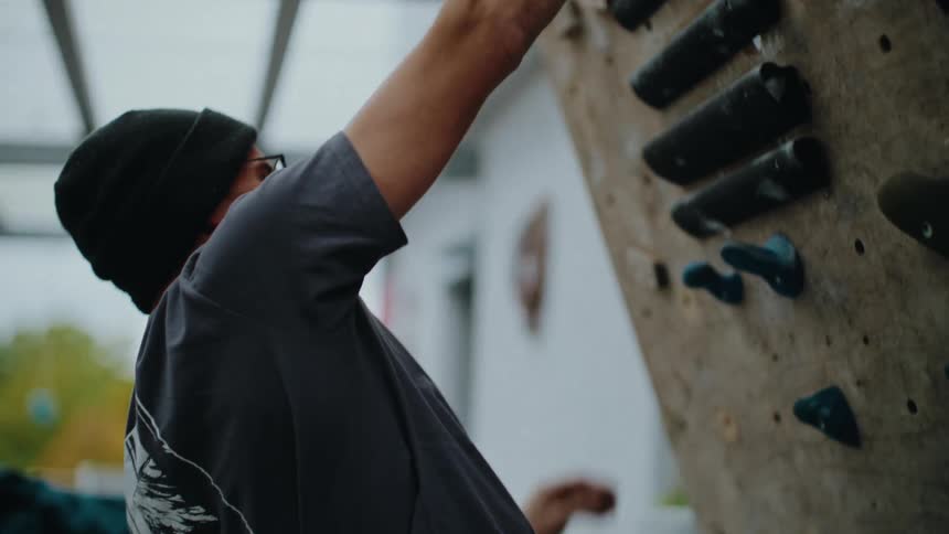 Slow motion close-up of a focused man in a beanie climbing up a wooden bouldering wall