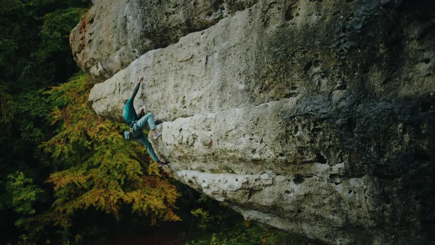 A strong rock climber tackles a difficult overhang on a forest cliff, eventually falling and being safely caught by his rope