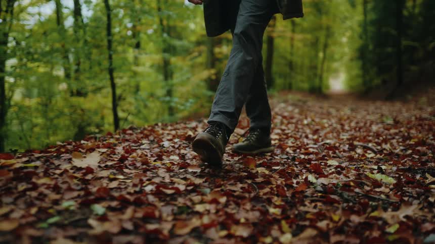 A slow-motion close-up shot focuses on a person's hiking boots walking through a dense layer of fallen leaves on a forest path during autumn