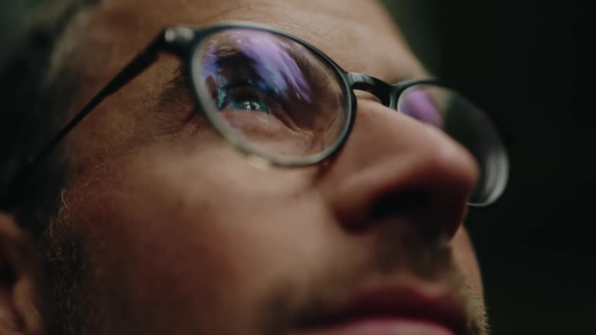 An extreme close-up of a contemplative man opening his eyes in slow motion, with his surroundings reflected in his glasses