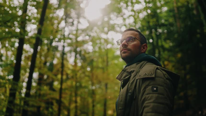 A thoughtful man wearing glasses stands peacefully in a tranquil forest, looking up at the sunlit canopy of tall trees