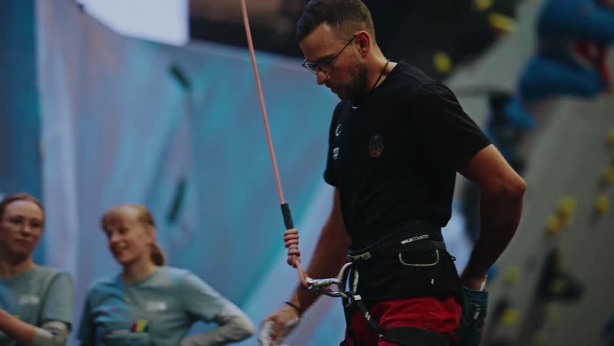 A focused male rock climber with glasses applies chalk to his hands and prepares to begin his ascent on an indoor climbing wall