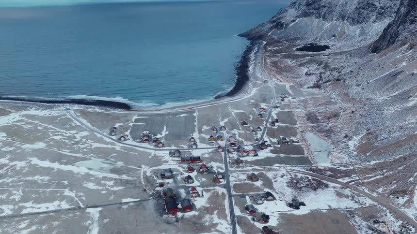 A stunning aerial view of a remote Scandinavian coastal village covered in snow, nestled between the calm ocean and a massive mountain