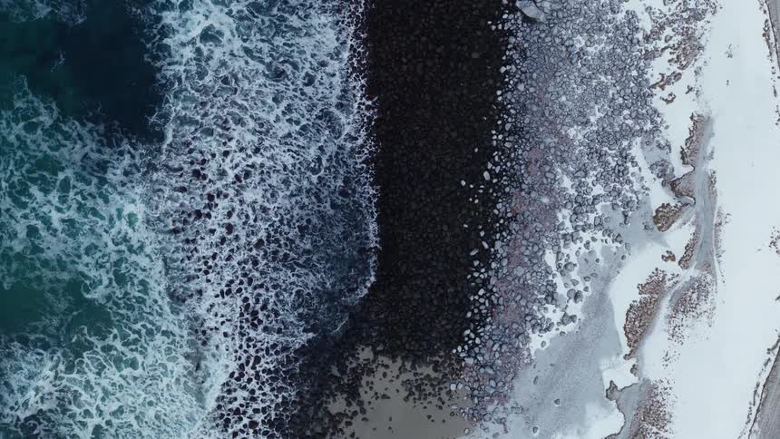 A slow, descending top-down aerial view of a dramatic, snowy coastline where frigid turquoise waves crash against the rocky shore