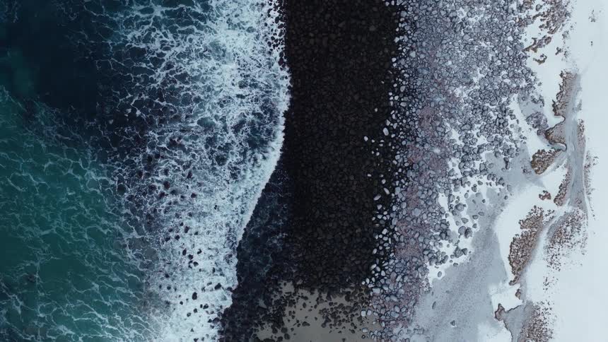 An ascending top-down aerial shot captures the contrast of dark ocean waves washing over a rocky, snow-covered beach