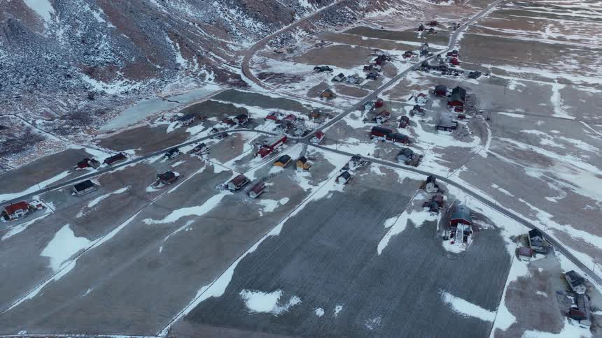 High-angle aerial view of a small, remote village nestled in a snowy valley at the base of a mountain