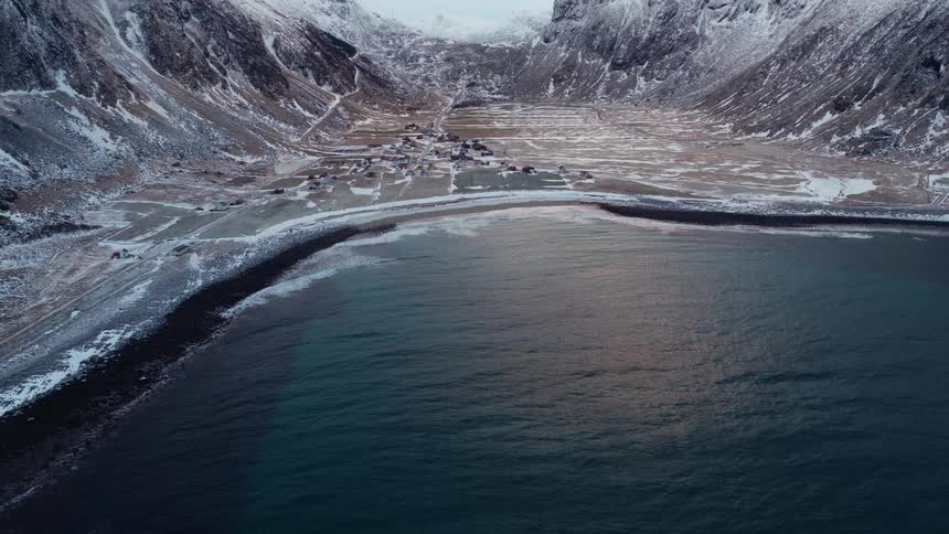 A breathtaking aerial shot reveals a secluded coastal village nestled between majestic, snow-capped mountains and the tranquil sea during winter
