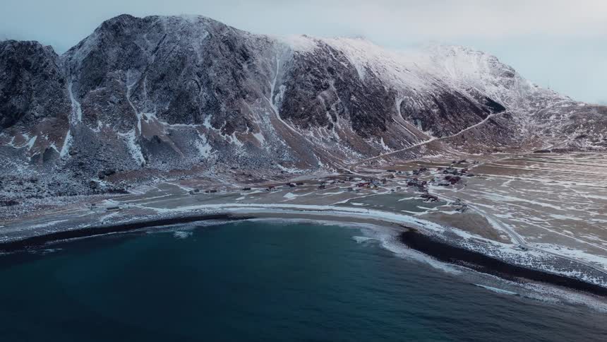 A serene aerial view shows a remote coastal village nestled in a snowy bay at the foot of majestic mountains