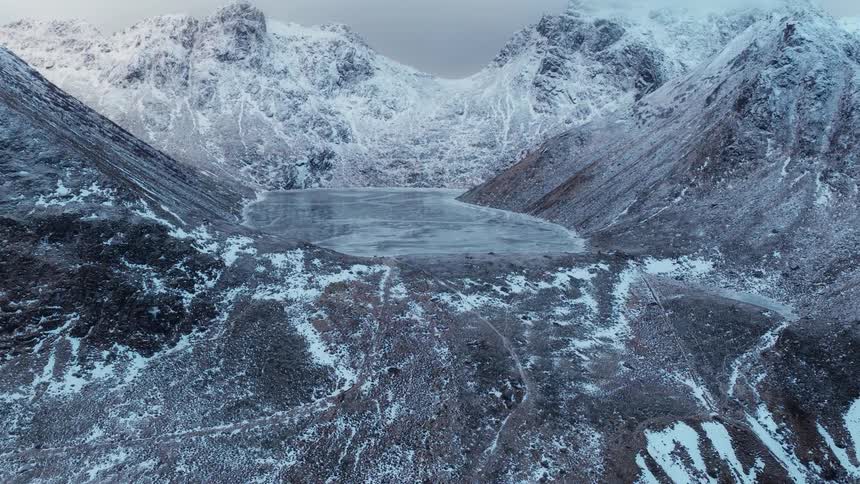 An epic aerial shot pushes forward over a vast, snowy landscape towards a frozen mountain lake