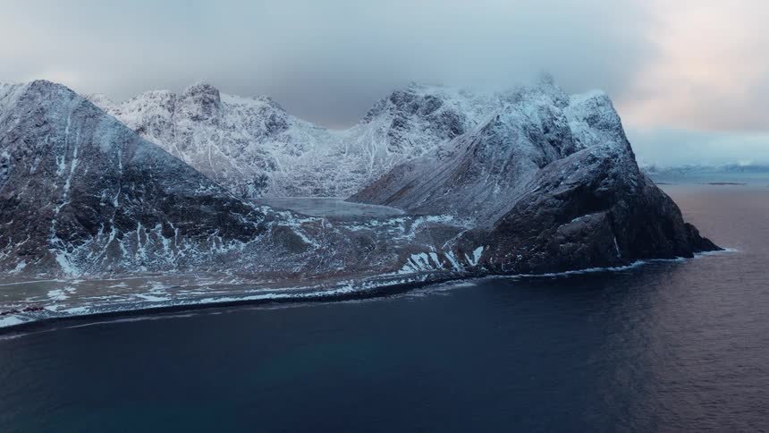 A majestic and dramatic aerial view of snow-covered mountains meeting the dark sea on a cold winter day in Norway